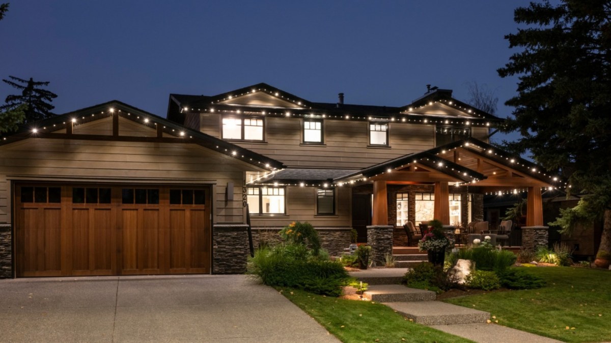 A 2 storey home and garage with multiple roof lines with a combination of Gemstone regular and flood light style white lights on. Around the yard are multiple flood lights illuminated throughout the greenery. Above the picture are the words, On the Blog, the difference between uplighting and downlighting with flood lights.